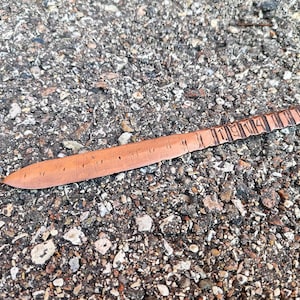 Puede incluir: Una regla de metal delgada de color cobre con una punta puntiaguda y los números 11 a 14 grabados en la superficie. La regla está sobre una superficie rugosa y gris.