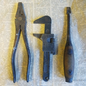 May include: A selection of antique tools, including pliers, a spanner, and a screwdriver, displayed on a newspaper backdrop. The tools show signs of wear and tear, suggesting they are for mechanical or repair tasks.
