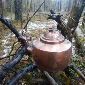 Könnte beinhalten: Ein Kupferkessel hängt an einem Ast in einer Waldlandschaft. Der Kessel ist alt und hat eine Patina des Alters. Der Waldboden ist mit Schnee und Blättern bedeckt.