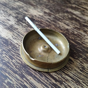 May include: A vintage brass ashtray with a cigarette inside. The ashtray is round with a small circular base and a slightly raised rim. The surface of the ashtray shows signs of wear and age. The background is a wooden surface.