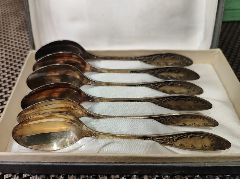 May include: Set of six silver teaspoons with ornate handles in a white box. The spoons are arranged in a row, with the handles facing the viewer.
