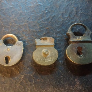 May include: Three antique brass padlocks with keyholes. The padlocks are of different sizes and shapes. The padlocks are on a brown leather surface.