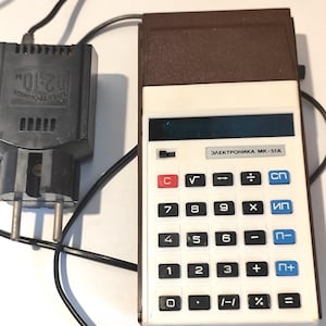 Könnte beinhalten: Ein brauner und weißer Taschenrechner mit einem schwarzen Stromkabel und einem schwarzen Netzteil. Der Rechner hat ein blaues Display und eine weiße Tastatur mit schwarzen Tasten. Die Tasten sind mit Zahlen, mathematischen Symbolen und dem Text "ЭЛЕКТРОНИКА МК-57А" beschriftet.
