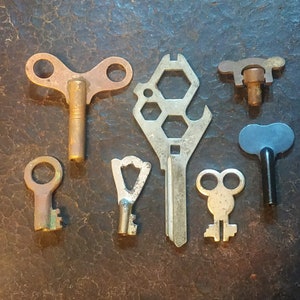 May include: A collection of seven vintage keys, including a multi-tool key with a hexagonal pattern, a key with two loops, and a key with a single loop. The keys are made of metal and have a worn, antique look.