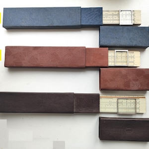 Puede incluir: Tres reglas de cálculo antiguas con sus estuches originales. Los estuches son azules, rojos y marrones. Las reglas de cálculo son beige con marcas en blanco y negro y un cursor transparente. Se utilizan para cálculos matemáticos.