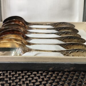 May include: A set of six silver spoons with ornate handles, displayed in a wooden box. The spoons are arranged in a row, with the handles facing the viewer.