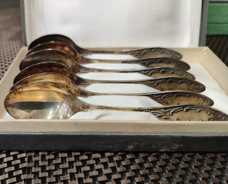 May include: A set of six silver spoons with ornate handles, displayed in a wooden box. The spoons are arranged in a row, with the handles facing the viewer.