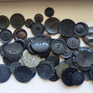 Puede incluir: Una colección de botones negros con varios diseños y texturas. Los botones están dispuestos en un patrón aleatorio sobre una superficie blanca.