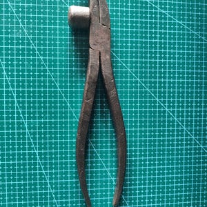 Op de afbeelding: Een vintage metalen nijptang met een donkere, verweerde afwerking, een gebogen bekontwerp en een kleine cilindrische uitstulping aan de zijkant. Het gereedschap vertoont zichtbare gebruikssporen, met het cijfer 14 in het metaal gestanst.