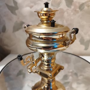 May include: A gold-coloured samovar with black accents and a spout. The samovar is sitting on a reflective surface.