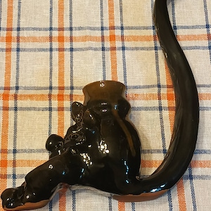 May include: A black ceramic pipe shaped like a seated figure with a long, curved stem. The figure has a round head and a long, flowing tail.
