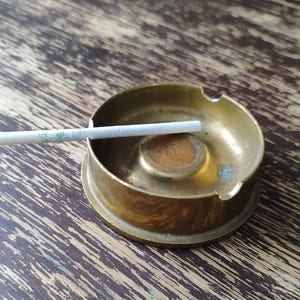 Puede incluir: Un cenicero redondo de latón con un cigarrillo apoyado en el borde. El cenicero tiene un diseño circular con un centro elevado y una pequeña muesca para sujetar el cigarrillo. El latón muestra signos de uso.