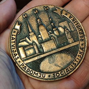 May include: A bronze coin with a depiction of a city skyline and the text "LATVIJAS PADOMJU SOCIALISTISKA REPUBLIKA" around the edge.