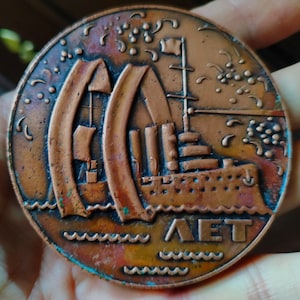 May include: A copper-colored medal with a detailed image of a ship sailing on the water. The ship has a tall mast and a large sail. The medal has a wavy border and the word "LET" inscribed on the bottom.