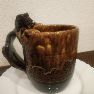 May include: A brown ceramic mug with a textured glaze and a unique handle shaped like a small animal. The mug has a brown and yellow colour scheme.