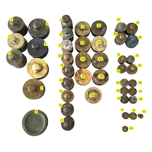 Puede incluir: Una colección de pesas antiguas de latón y bronce de varios tamaños y formas, incluyendo diseños cilíndricos, de disco y de campana. Algunas pesas tienen marcas visibles. La colección está dispuesta sobre una superficie blanca.