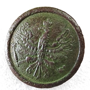 Puede incluir: Un botón de metal verde con una imagen en relieve de un águila coronada. El águila tiene un diseño detallado con alas extendidas y un pico afilado.