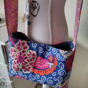 Könnte beinhalten: Eine blaue Stoff-Umhängetasche mit einem farbenfrohen Blumenmuster in Rosa, Orange und Beige. Die Tasche hat einen rosa Riemen mit einem floralen Design. Die Tasche wird auf einer weißen Schaufensterpuppe präsentiert.