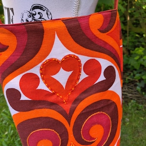Könnte beinhalten: Eine farbenfrohe Umhängetasche mit Retro-Design. Die Tasche hat einen weißen Hintergrund mit wirbelnden Mustern in Orange, Braun und Rot. Ein herzförmiges Design, verziert mit orangefarbenen Perlen, ist mittig auf der Vorderseite angebracht. Sie hat einen roten Riemen.