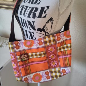 Könnte beinhalten: Schultertasche mit Retro-Blumen- und Karomuster in Orange, Braun, Rosa und Weiß. Die Tasche hat einen schwarzen Riemen und einen kleinen gestickten Aufnäher. Der Text "PURE NATURE FASHION LIFE" ist auf der Schaufensterpuppe zu sehen.