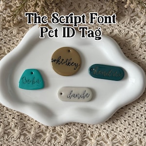 May include: A white ceramic tray holds four pet ID tags in various shapes and colors. The tags are teal, tan, and white, with script font names: "Boba", "whiskey", "hunter", and "dande". The text "The Script Font Pet ID Tag" is above the tags.