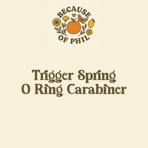 May include: Cream-colored graphic with the text "BECAUSE OF PHIL" above the words "Trigger Spring O Ring Carabiner." The top graphic includes an orange, butterfly, lemon, and flowers.