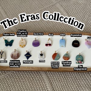 May include: The Eras Collection presents a selection of colourful charms. The charms include a butterfly, a jacket, a forest scene, a diamond, a bow, a heart, and other shapes. The charms are displayed on a wooden tray.