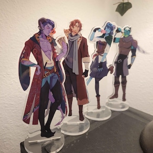 May include: Four acrylic character standees featuring fantasy figures. The characters are in various poses, with detailed clothing and accessories. Each standee is mounted on a clear, circular base. The color palette includes shades of purple, brown, blue, and green.