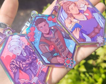May include: Three iridescent acrylic keychains with fantasy character illustrations. Each keychain is a different character, with vibrant colors and floral accents. The keychains are hexagonal and have silver-colored hardware.