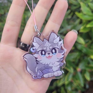 May include: A clear acrylic charm featuring a cartoon wolf with gray fur, pink cheeks, and blue eyes. The charm has a pink string attached for hanging. The wolf is smiling and has a blue band around its leg.