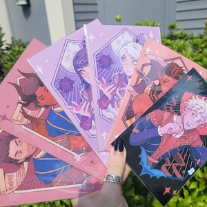 Könnte beinhalten: Eine Hand hält einen Fächer aus Fantasy-Kunstdrucken. Die Drucke zeigen Illustrationen von Charakteren in verschiedenen Farben, darunter Rosa, Lila und Blau. Das Kunstwerk ist vor einem Hintergrund aus grünem Laub und einem blauen Gebäude platziert.