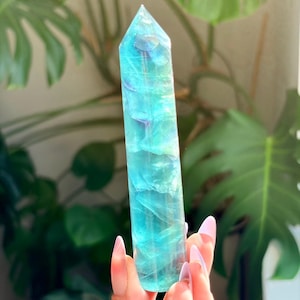 May include: A polished, hexagonal fluorite crystal tower featuring translucent layers of vibrant teal, blue, and subtle purple hues. The stone has a smooth, faceted surface that catches the light, revealing internal fractures and natural color variations.