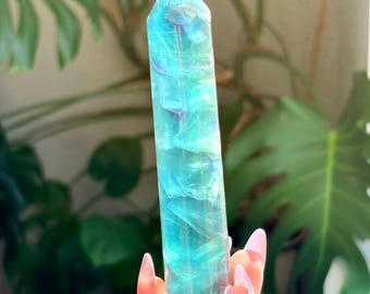 May include: A polished, hexagonal fluorite crystal tower featuring translucent layers of vibrant teal, blue, and subtle purple hues. The stone has a smooth, faceted surface that catches the light, revealing internal fractures and natural color variations.