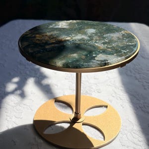 May include: A circular display stand featuring a polished moss agate stone top with green, white, and brown marbled patterns. The stand has a gold-coloured metal frame with a slender central post and a flat, round base with four petal-shaped cutouts.