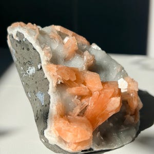 Könnte beinhalten: Ein Mineralexemplar mit einer Ansammlung durchscheinender, pfirsichfarbener Kristalle, die aus einer grauen, texturierten Gesteinsbasis wachsen. Die Kristallstruktur zeigt scharfe, kantige Formationen mit glänzendem Finish, die einen Kontrast zur matten, dunkelgrauen Außenseite des Steins bilden.