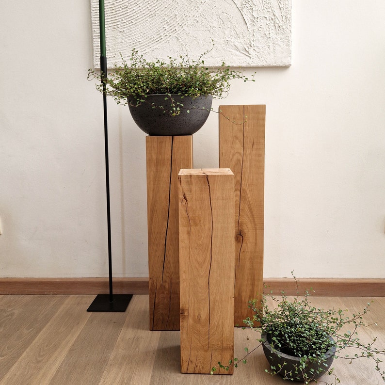 May include: Three wooden plant stands of varying heights, made from natural wood with visible grain. The stands are arranged in a row, with a small potted plant on the tallest stand and a larger potted plant on the middle stand.