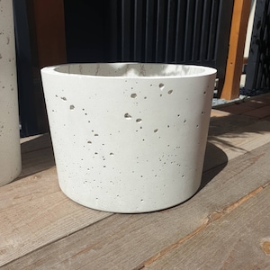 Könnte beinhalten: Ein weißer Betontopf mit gesprenkelter Oberfläche. Der Topf ist zylindrisch und hat einen glatten, abgerundeten Rand.