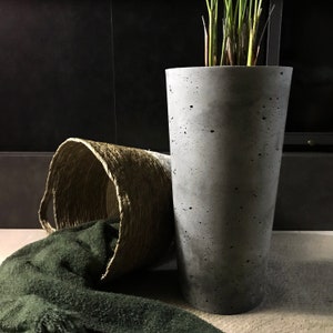 Könnte beinhalten: Ein grauer Betontopf mit rauer Textur, der auf einer beigefarbenen Oberfläche steht. Im Hintergrund befinden sich ein geflochtener Korb und eine grüne Decke.