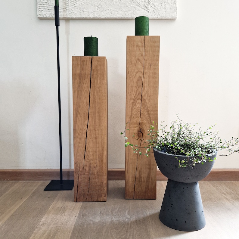 May include: Two wooden candle holders with a natural wood finish and a crack running down the center of each. The candle holders are on a light wood floor. A small potted plant is in the foreground.