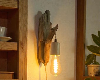 May include: A rustic wall sconce featuring a piece of driftwood with a light bulb hanging from it. The light bulb is a warm yellow color, and the driftwood is a light brown color. The sconce is mounted on a white wall.