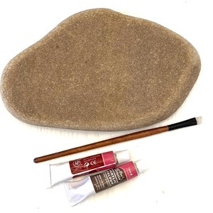 Pode incluir: Uma pedra castanha lisa de forma irregular, um pincel de madeira e dois tubos de tinta acrílica. Um tubo é branco com tinta vermelha, e o outro é branco com tinta castanha. Os tubos de tinta estão rotulados como "Acrylic Colour".