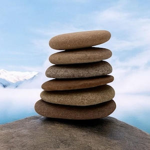 Può includere: Un cumulo di sei pietre lisce e marroni, bilanciate l'una sull'altra, su una grande roccia grigia. Le pietre sono impilate a forma di piramide, con la più grande in basso e la più piccola in alto. Lo sfondo è un cielo blu con nuvole bianche e un'estensione d'acqua.