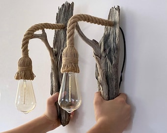 Puede incluir: Un aplique de pared que presenta dos piezas de madera flotante gris desgastada. Cada pieza sostiene un cordón de cuerda trenzada de color tostado que cuelga hacia abajo, terminando en un casquillo envuelto en cuerda con una bombilla clara.