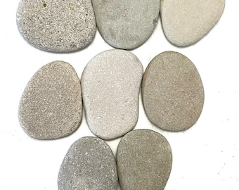 May include: A collection of seven smooth, rounded river stones arranged in a cluster. The stones vary in color, featuring shades of light gray, beige, tan, and speckled granite, each with a unique, naturally weathered texture and irregular shape.