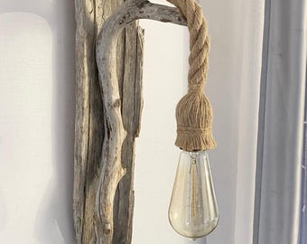 Puede incluir: Un aplique de pared con una placa de madera flotante gris desgastada y un brazo de rama curvada. Una cuerda de yute gruesa de color natural se extiende desde la parte superior, sosteniendo una bombilla Edison transparente en forma de pera.