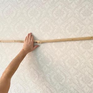 Op de afbeelding: Een lichtbruine houten stok, horizontaal gehouden tegen een behang met patroon. De stok is lang en slank, met een natuurlijk, onbewerkt uiterlijk. De hand die de stok vasthoudt is zichtbaar.