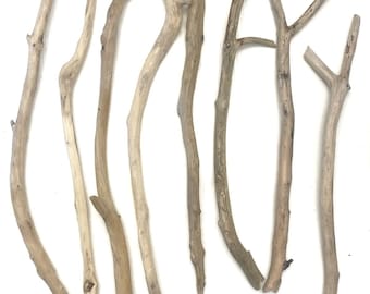 Peut inclure: Huit morceaux de bois flotté de différentes longueurs et formes, disposés sur un fond blanc. Les branches présentent des tons de bois naturels, allant du beige clair au brun plus foncé, avec des nœuds et des textures visibles.