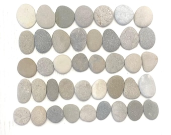 May include: A collection of smooth, oval-shaped stones in varying shades of gray, beige, and white, arranged neatly on a white surface. The stones have a natural, weathered appearance, suggesting they are river rocks or similar. The arrangement is symmetrical.