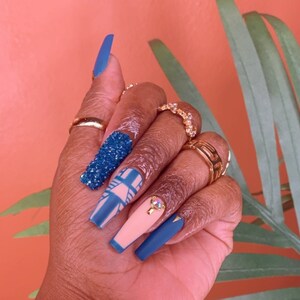 Peut inclure: Une main avec des ongles en gel bleu et beige. Les ongles sont décorés de paillettes, de motifs géométriques et de strass.