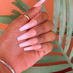 May include: Close-up of a hand with long, almond-shaped nails. The nails are painted with a white ombre design, transitioning from a light pink base to a white tip.